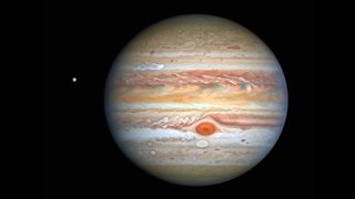 Image of Jupiter captured by the Hubble Space Telescope, showcasing the planet's swirling, colorful atmosphere. Prominent bands of clouds in shades of orange, brown, and white stretch across the gas giant's surface, with a distinct reddish-brown oval marking the Great Red Spot—a massive storm larger than Earth. A small moon shines to the left, this is Europa.