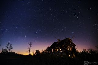 meteors streak through a star-studded sky with a house in the foreground.