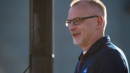 a man wearing a dark nasa logo polo shirt and glasses smiles with a greying goatee.